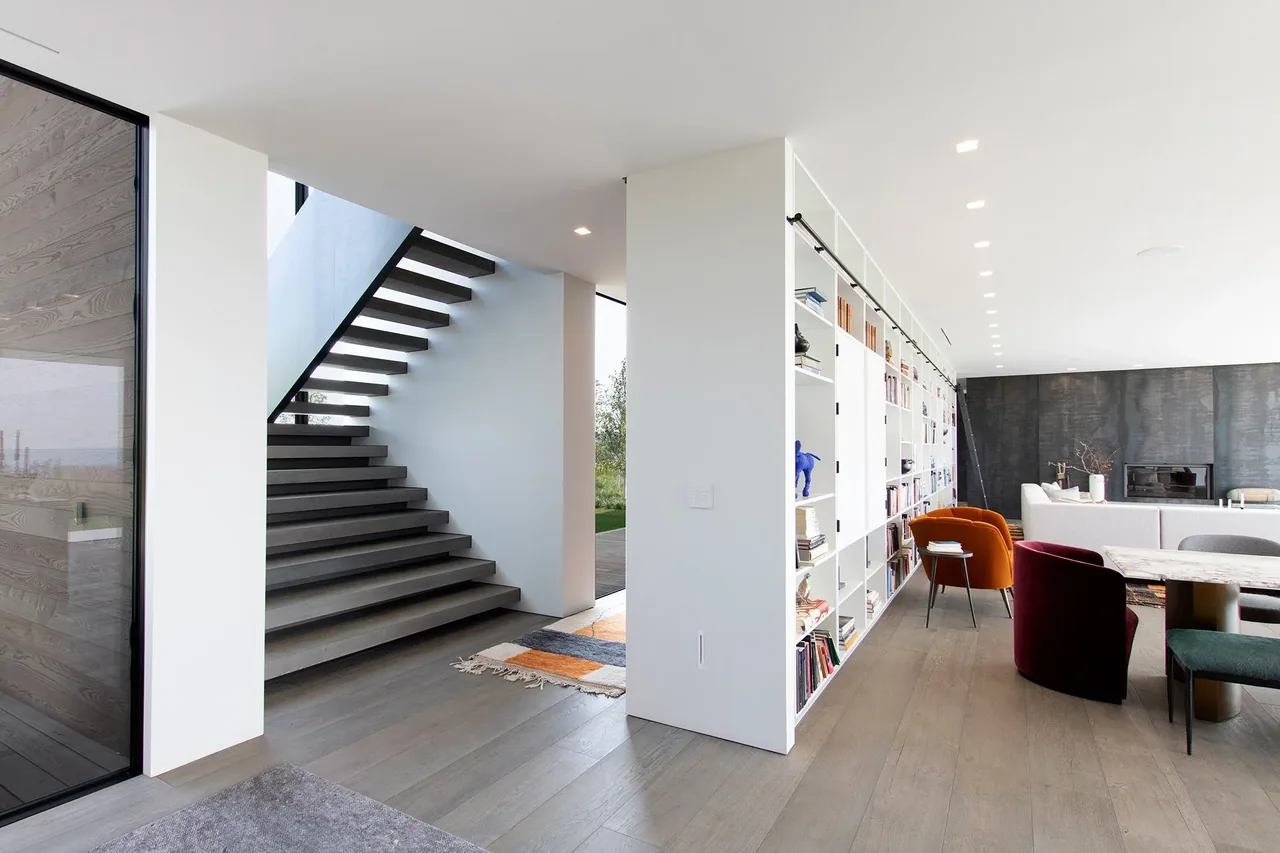 Millbrook Residence features floating wood stairs and an open living room with built-in white shelving and colorful chairs.