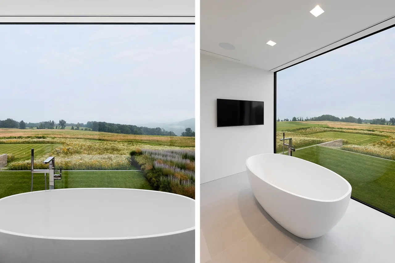 A modern bathroom features a white freestanding tub next to large corner windows with views of fields outside.