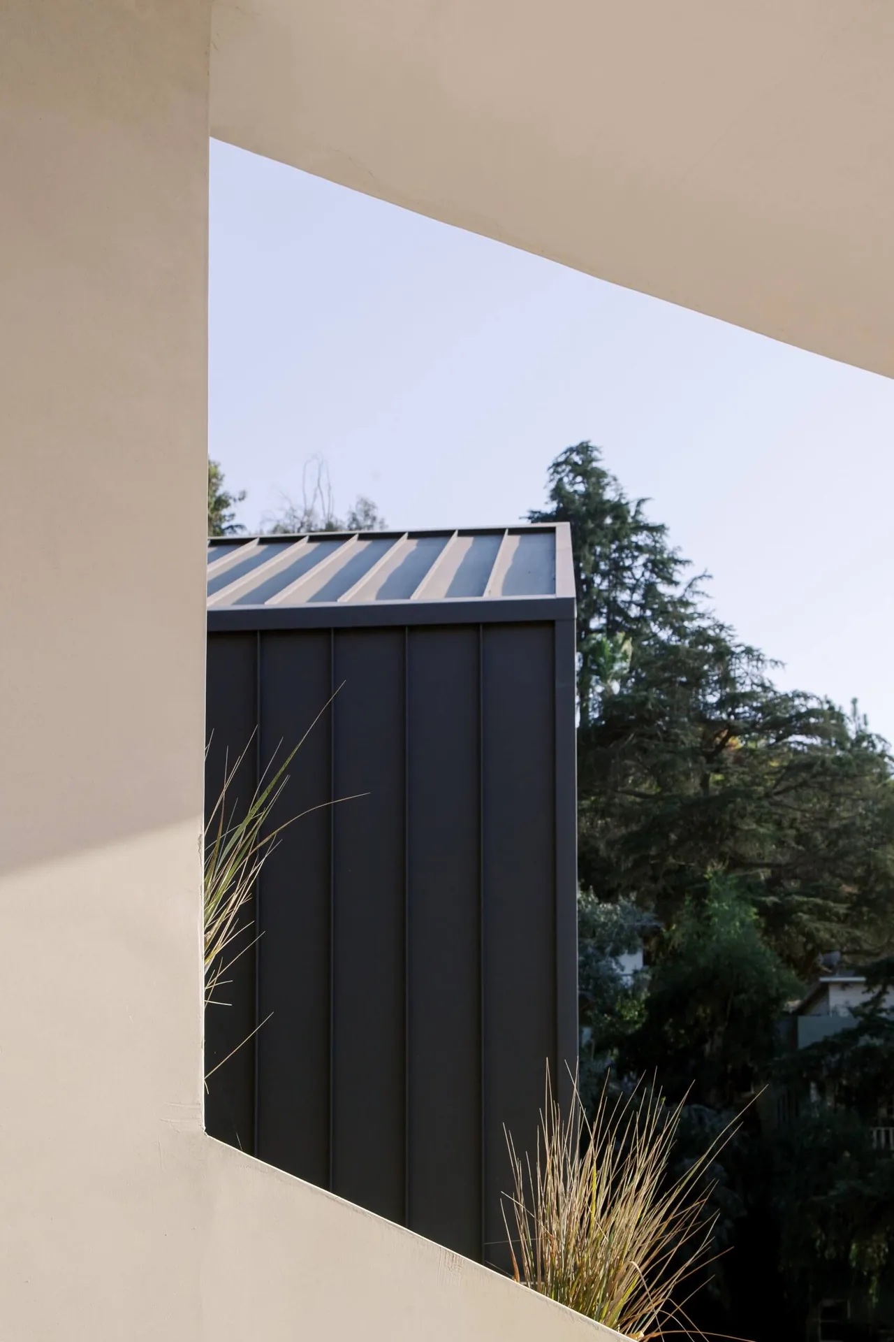 Metal standing seam roof with vertical panel siding visible through a stucco frame with foliage in the foreground.