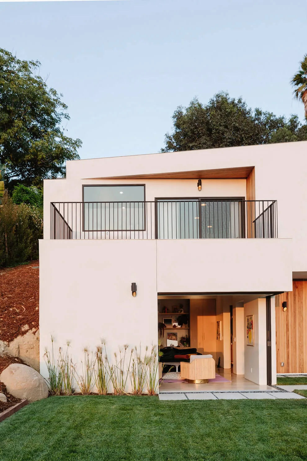 The Hillside Home + Studio features a modern white facade with a black metal railing balcony and a folding glass door leading to the outdoor grass patio.