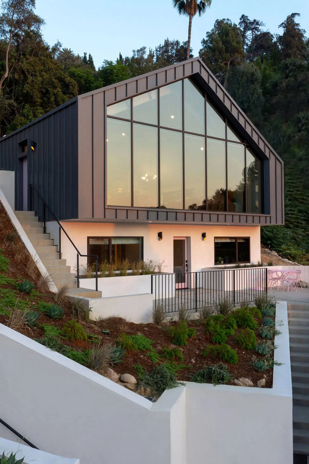 Modern hillside home featuring a large floor-to-ceiling window facade and black metal railings overlooking landscaped plantings.