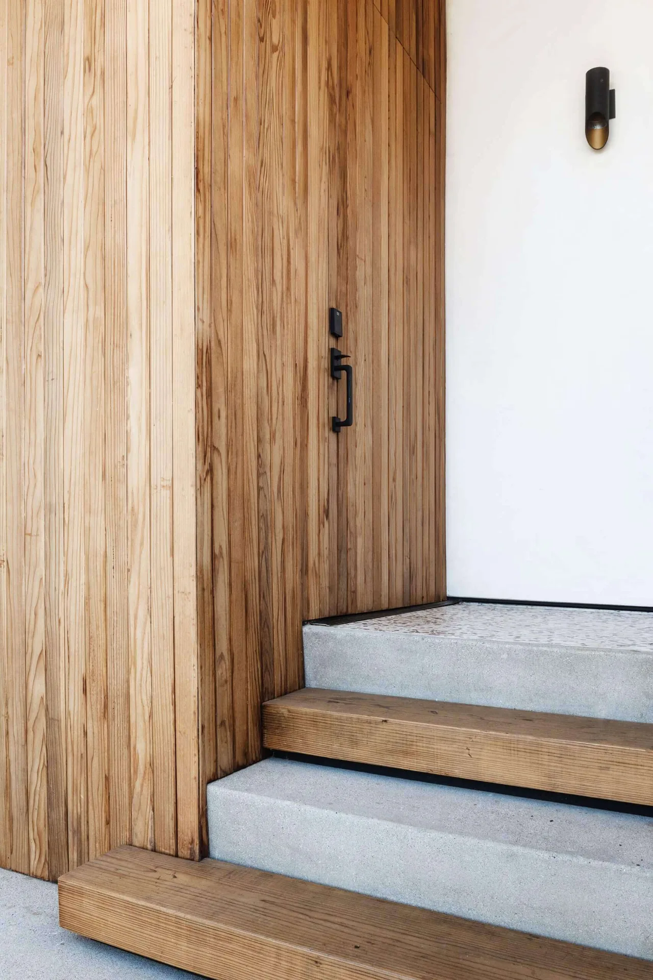 Entry area with vertical wood paneling, concrete steps with wood stair treads, and minimalist black wall sconce.