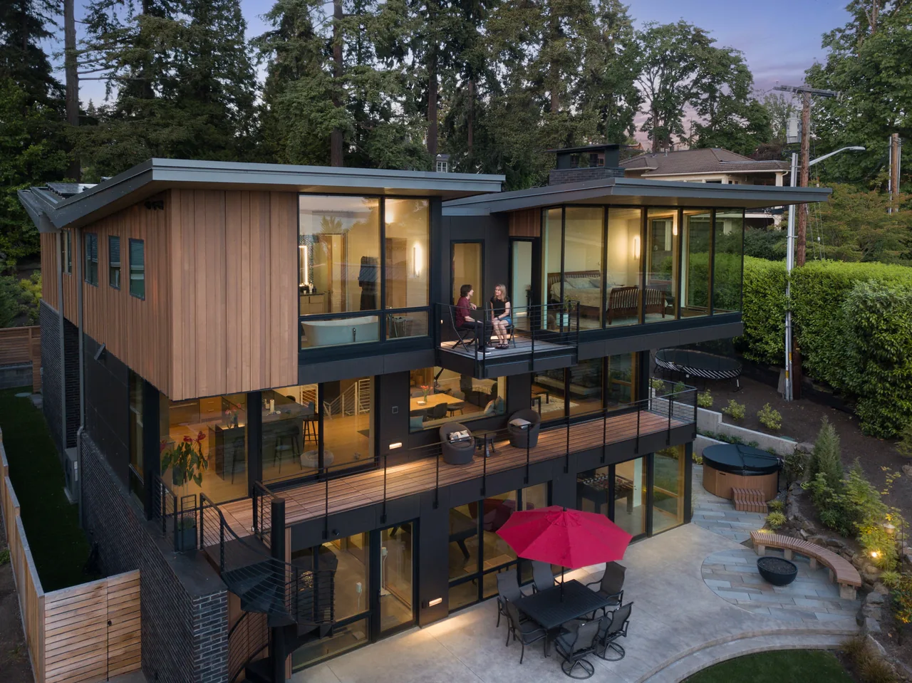 The Wingspan Residence features wood siding, extensive glazing, and outdoor terraces with metal railings and a red patio umbrella.