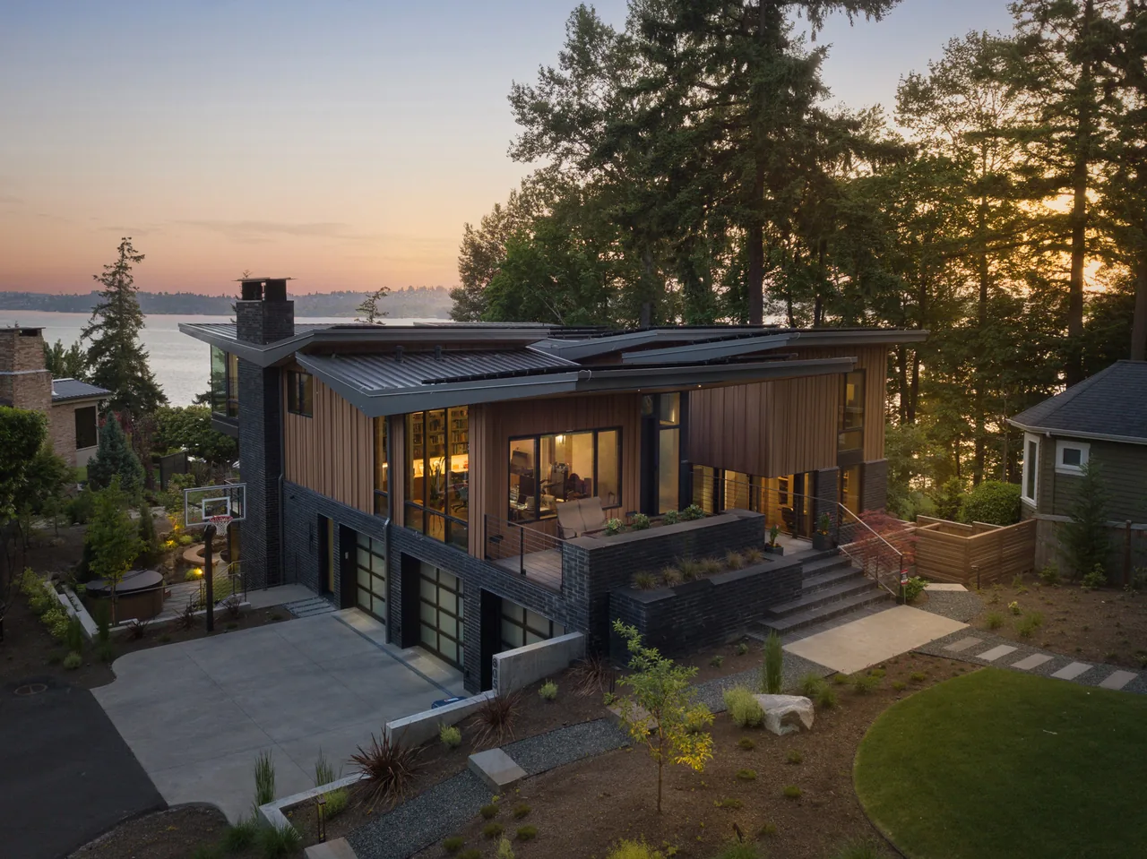 The Wingspan Residence features a metal roof, black brick lower level, and wood siding with expansive windows facing a forested waterfront.