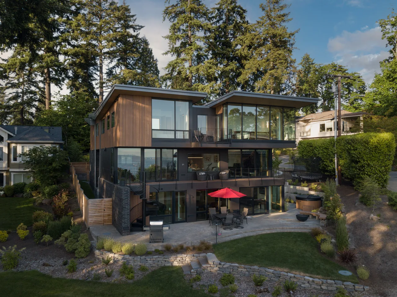 The Wingspan Residence features wood siding, floor-to-ceiling glazing, metal railings, and a red patio umbrella on the outdoor living area.