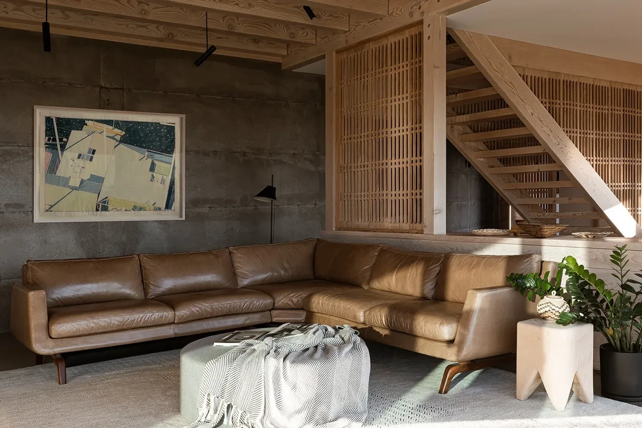 Mosier House living room features a tan leather sectional, wood slat divider, exposed wooden ceiling beams, and concrete wall.
