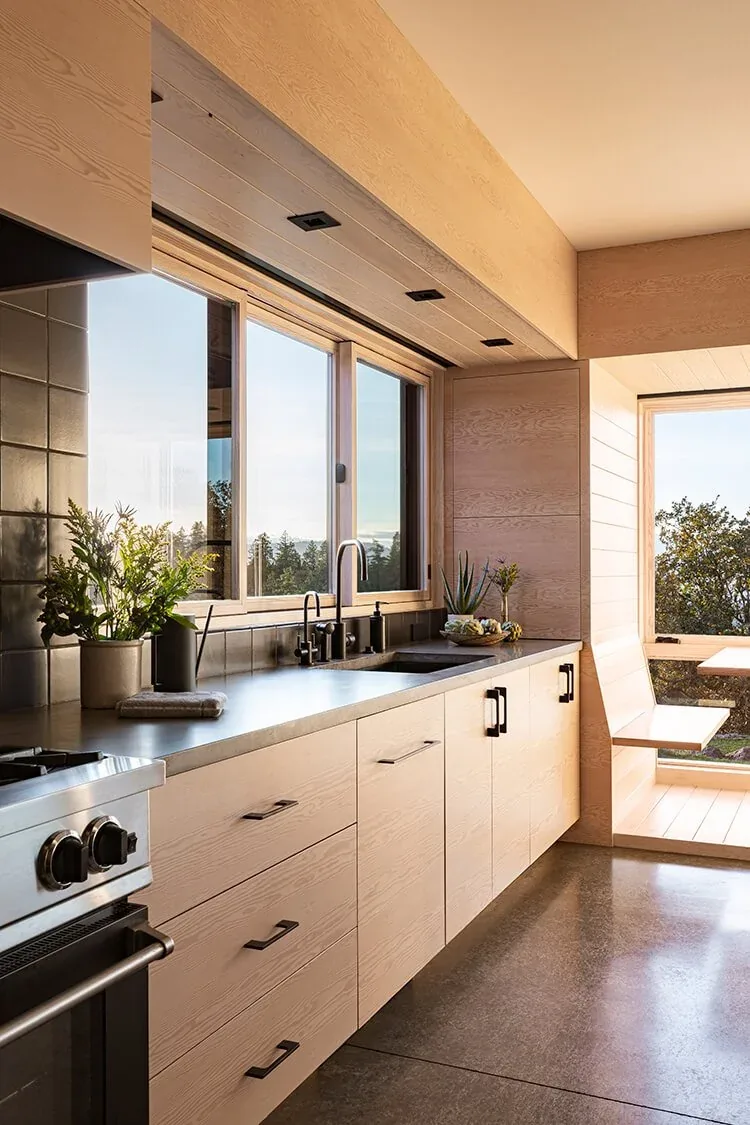 Kitchen with light wood cabinetry, black hardware, and large window offering a view of surrounding trees and hills.