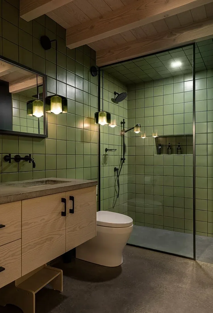 Bathroom in Mosier House features green tiled walls, wood ceiling beams, and a glass shower with black fixtures.