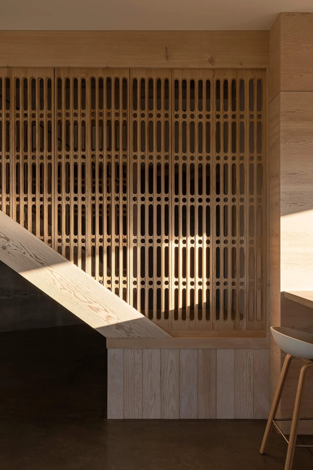 Wood slat room divider with patterned cutouts and light wood paneling under a staircase.