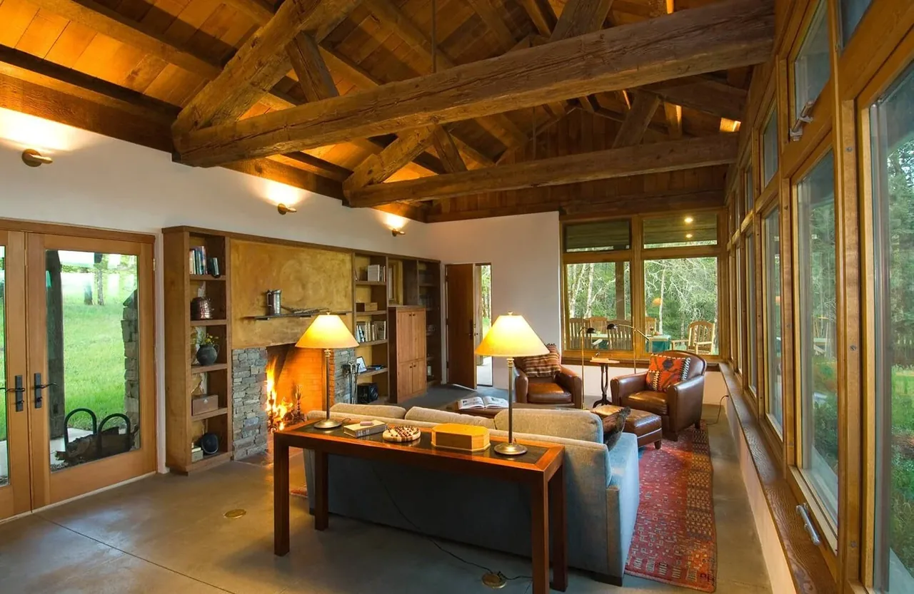 The living room of Columbia Gorge House features exposed wooden ceiling beams, a stone fireplace, and floor-to-ceiling windows overlooking a wooded lawn.