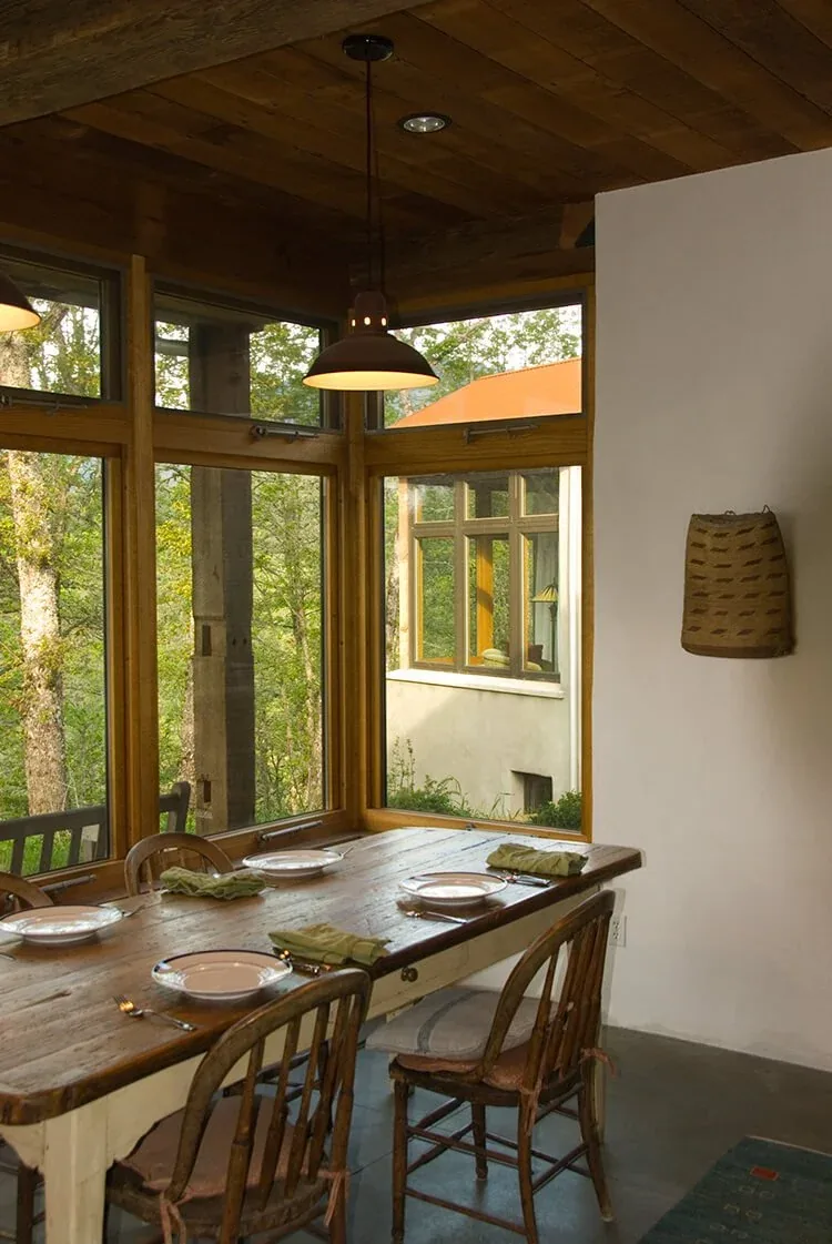 Dining room features a rustic wooden table, wood-framed windows, and a pendant light with views of wooded surroundings.