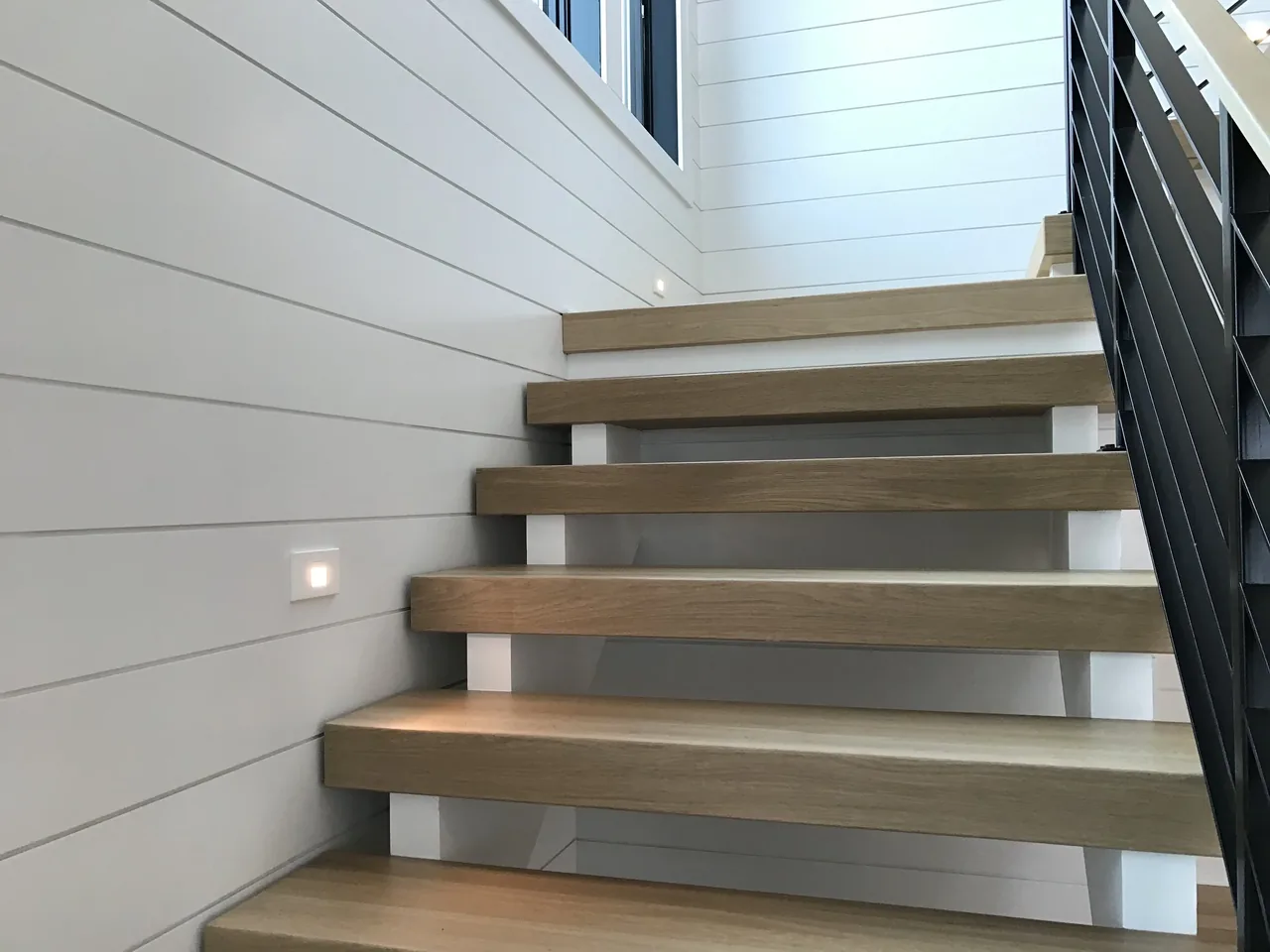Floating wooden staircase with white shiplap walls and black cable railing provides modern interior access.