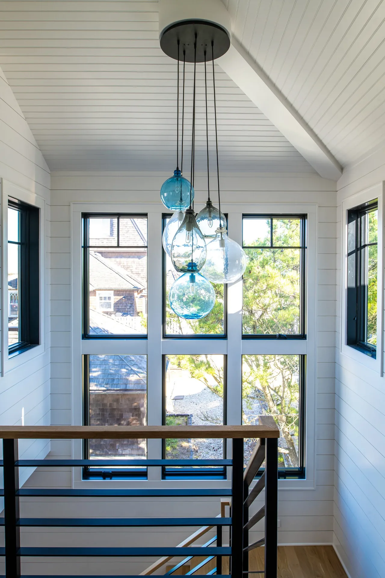 A group of glass pendant lights suspended in the vaulted ceiling stairwell with black window frames and white shiplap.
