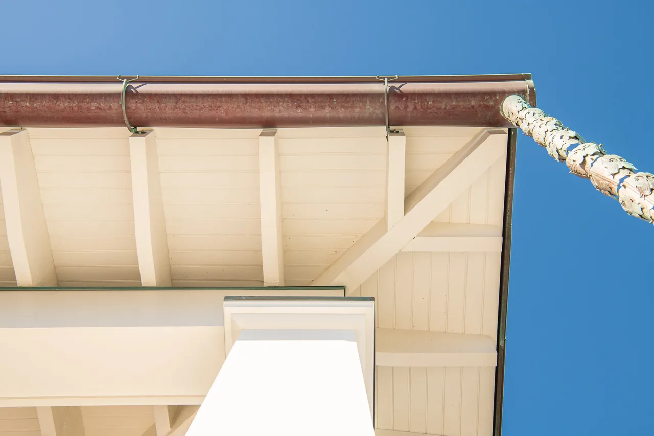Detail of Porch Talk House's white shiplap porch ceiling, tapered column, and rusted metal gutter under a blue sky.
