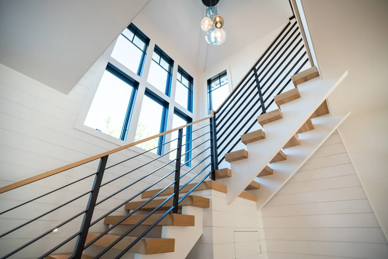 Interior staircase with floating wood treads and black metal railings beside large vertical black-framed windows within white shiplap walls.