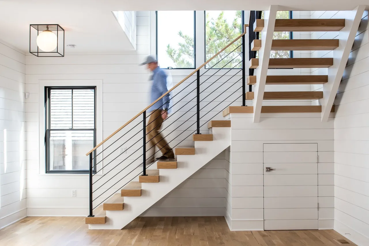 Modern interior staircase with wood treads and black metal railing, illuminated by a cube ceiling light and natural light from windows.