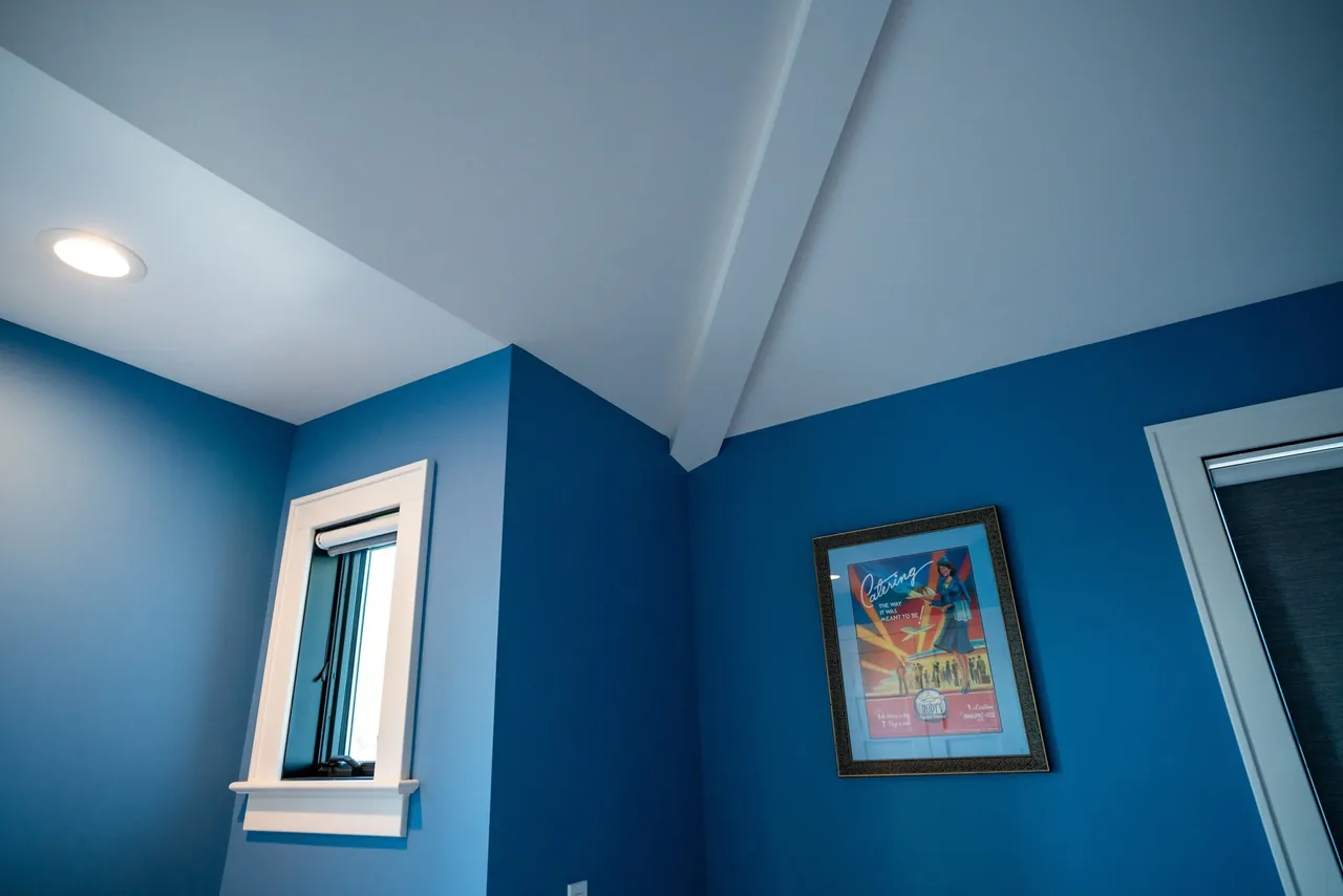 Blue interior corner with a small window, recessed ceiling light, and a colorful framed poster on the wall.