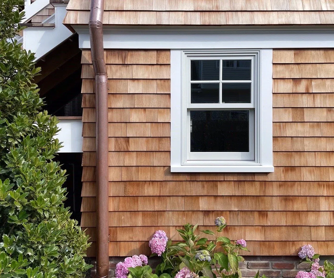 Window with divided lites set in white trim on a wood-shingle wall with a copper downspout beside it.