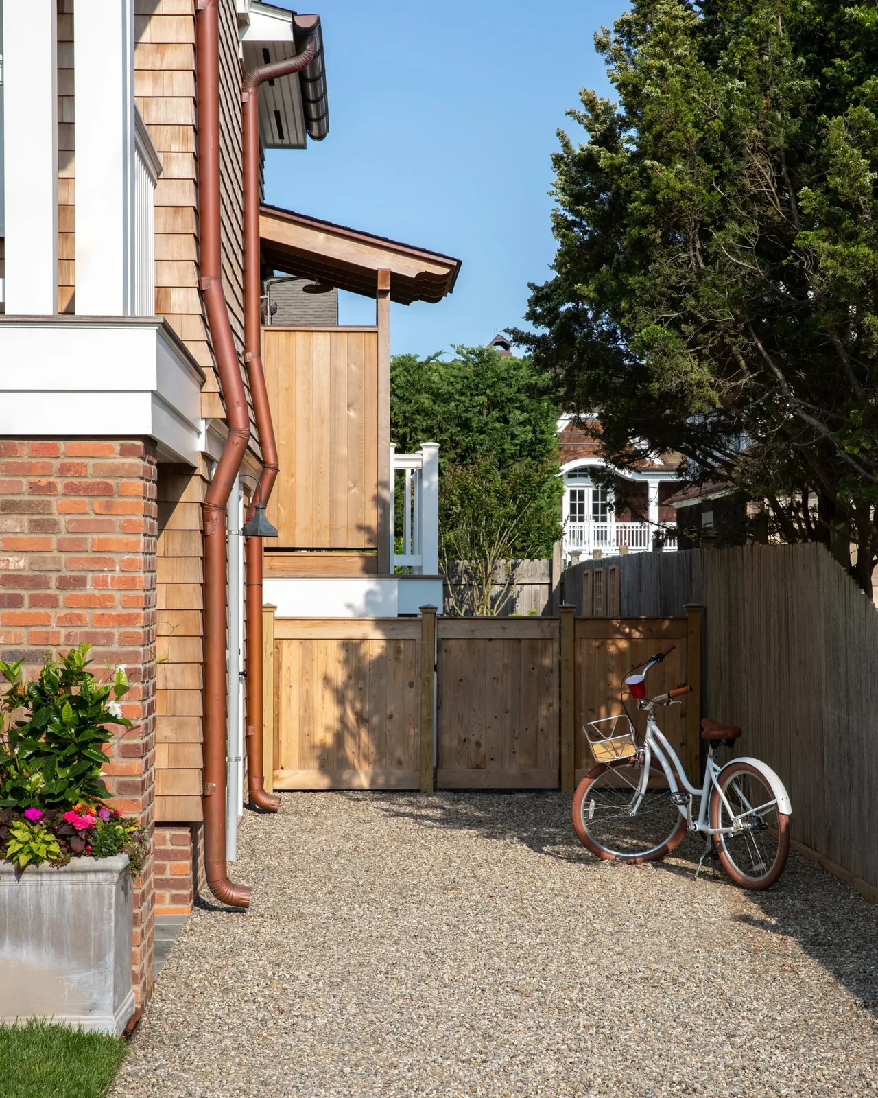 Side yard of a residence with brick and wood shingle exterior siding, a wooden gate, and a bicycle against the fence under clear sky.