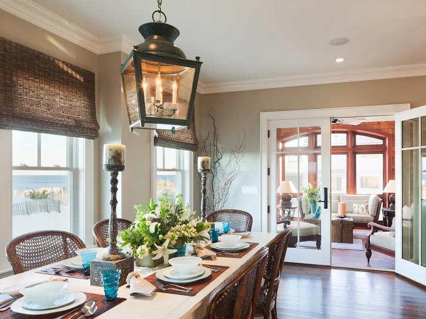 Dining room featuring a large white table with wicker chairs and a lantern-style pendant light, leading to a sunroom with wood trim.