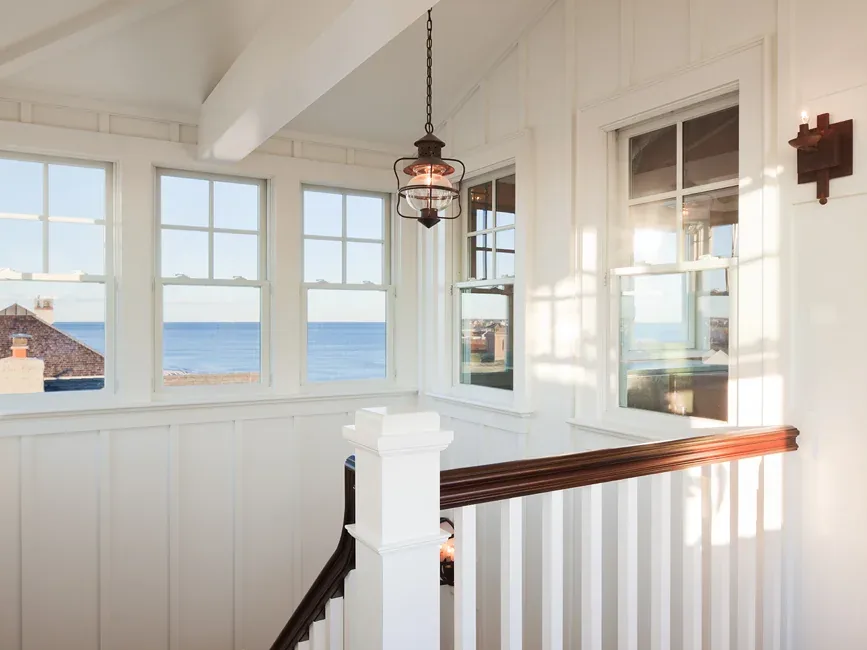 Stair landing with white walls, brown stair railing, multiple windows overlooking the ocean, and a hanging lantern pendant light.