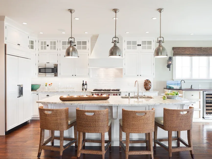 A bright kitchen features a large marble island with wicker bar stools and metal pendant lights by Richard Bubnowski Design.
