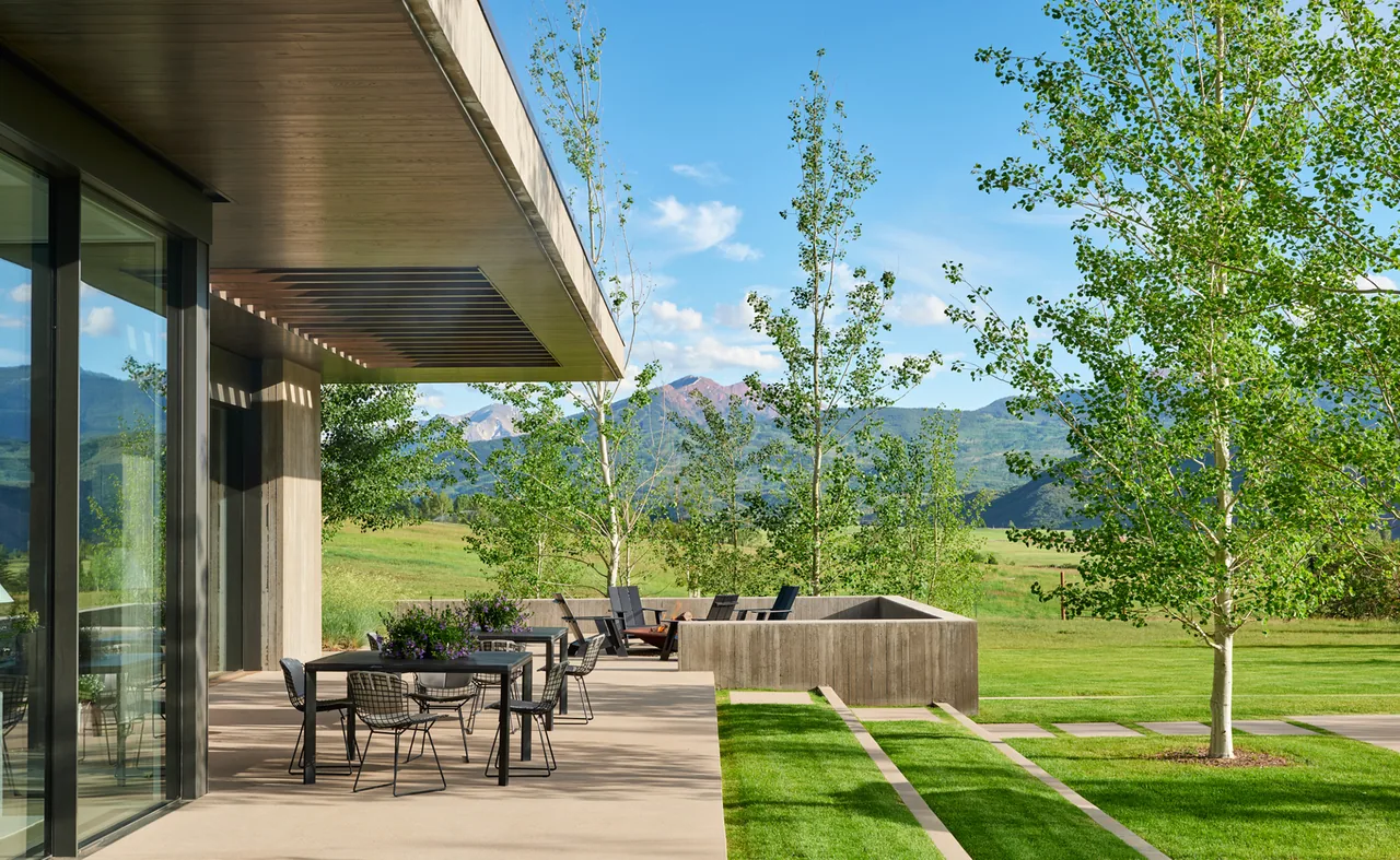 Modern outdoor patio featuring dining table, wire-frame chairs, and built-in seating overlooking mountains and grassy lawn.
