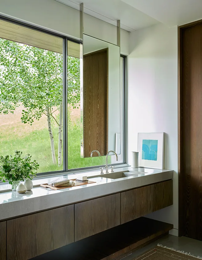 Bathroom sink with a large window looking out to greenery, featuring a hanging rectangular mirror and wood cabinetry.