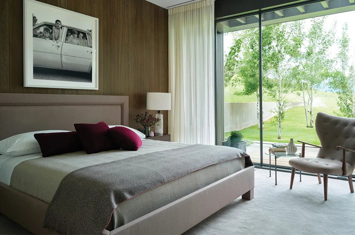 Modern bedroom with upholstered bed, wood-paneled wall, and sliding glass door revealing outdoor landscape.