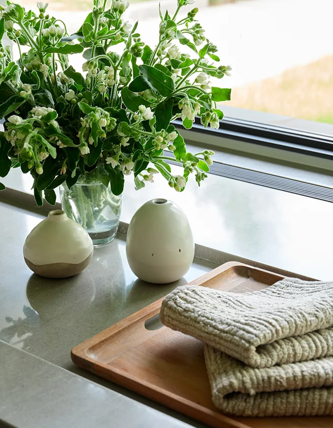 A kitchen counter features ceramic vases, fresh flowers, and folded towels near a slidable window.