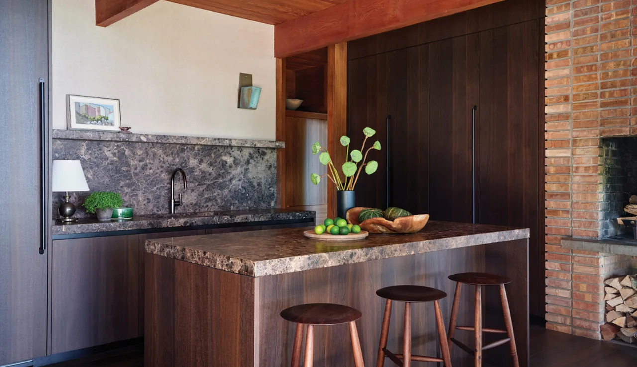 Midcentury kitchen featuring dark wood cabinetry, a stone countertop island, and a brick fireplace with wood storage.
