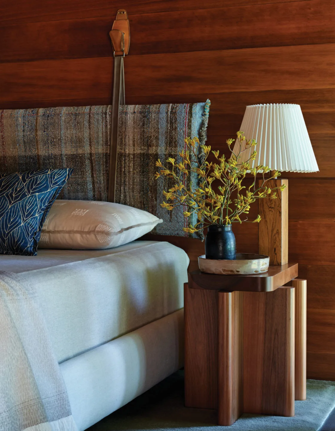 Bedroom corner features a wood-paneled wall, upholstered bed, and wooden side table with a lamp and floral arrangement.