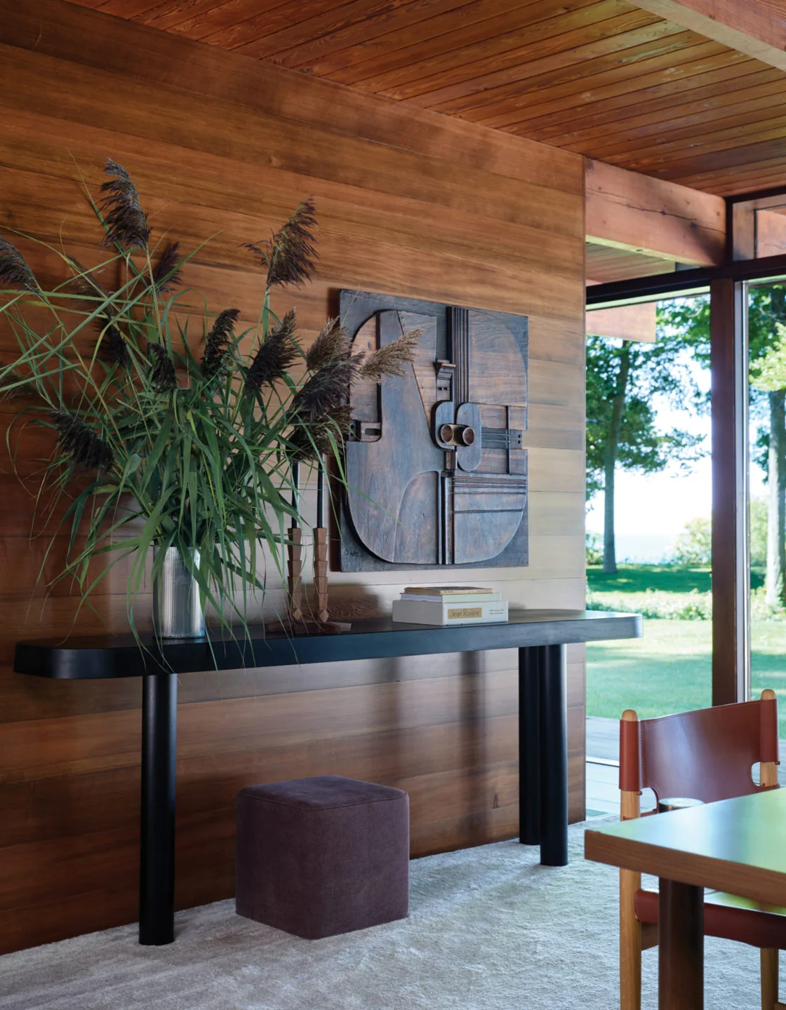 A dark console table with decor sits against a wood-paneled wall in a room with a large window view of the outdoors.