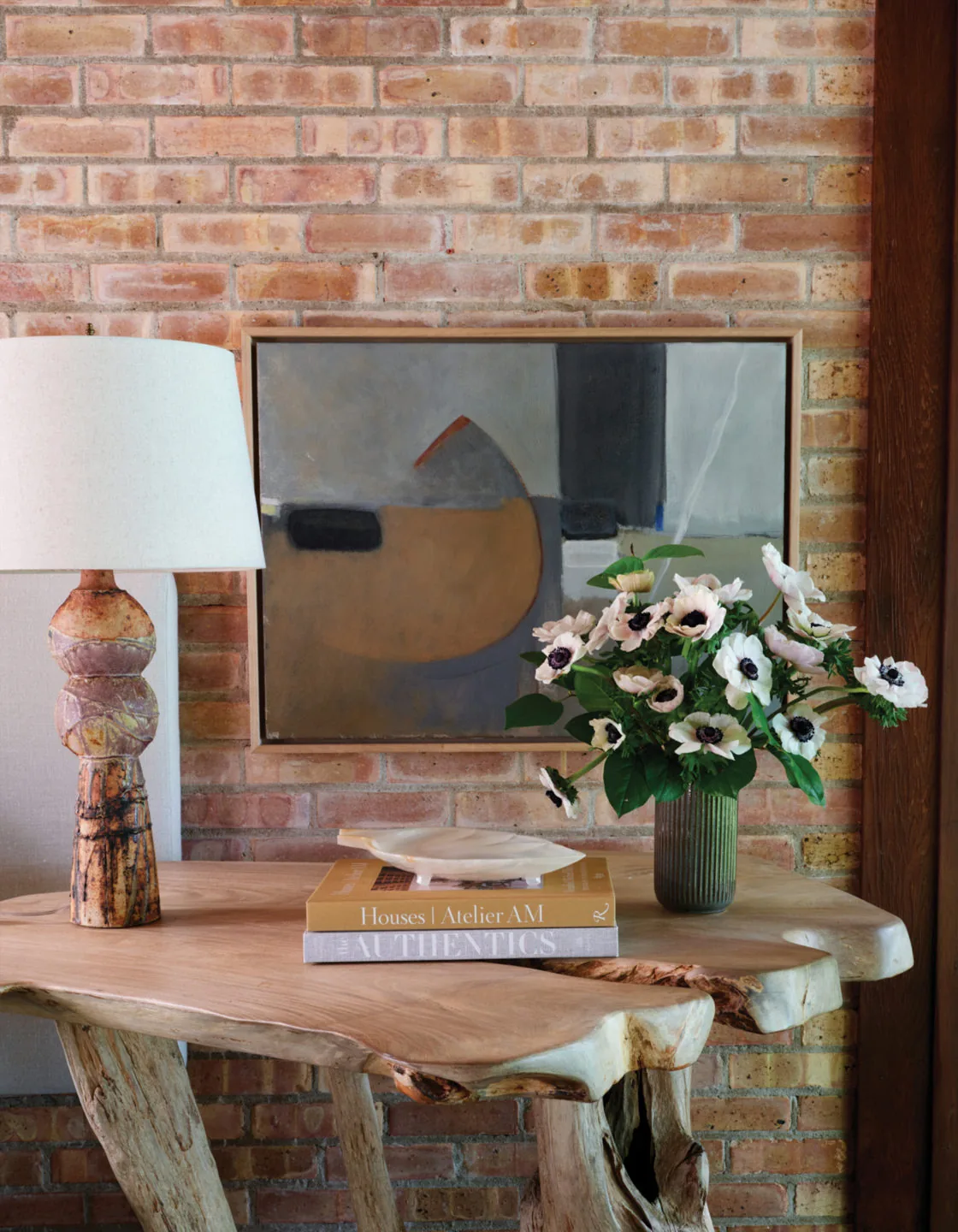 Rustic wooden table with a ceramic lamp, abstract wall art, and a floral arrangement in a brick-walled interior.