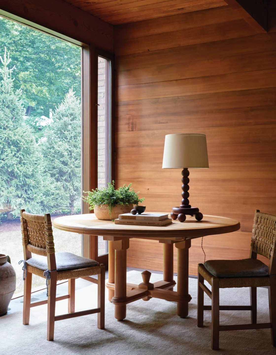 Wood-paneled dining nook with natural wood round table and woven chairs overlooking greenery through a large window.