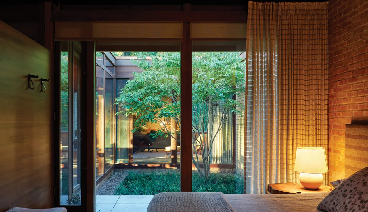 The bedroom overlooks a green courtyard through floor-to-ceiling glazing with wood framing and brick wall accents.