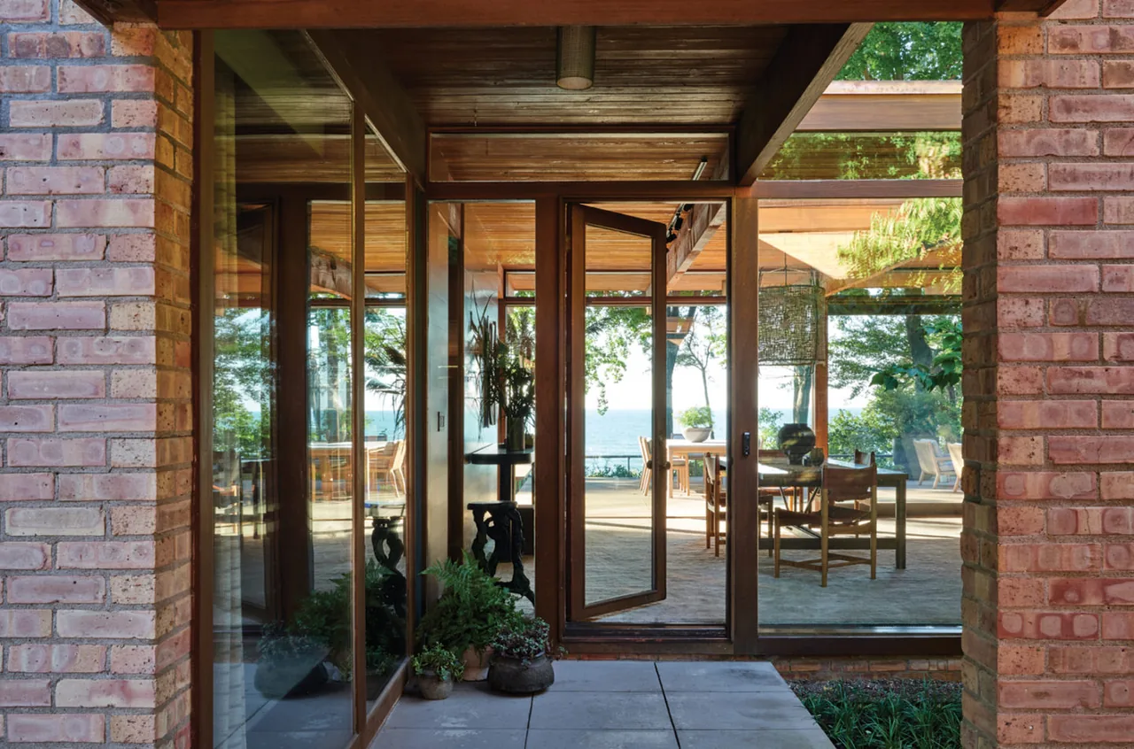 Lakeside Midcentury entry with wood framed glass pivot door and brick walls leading to a patio with dining furniture.