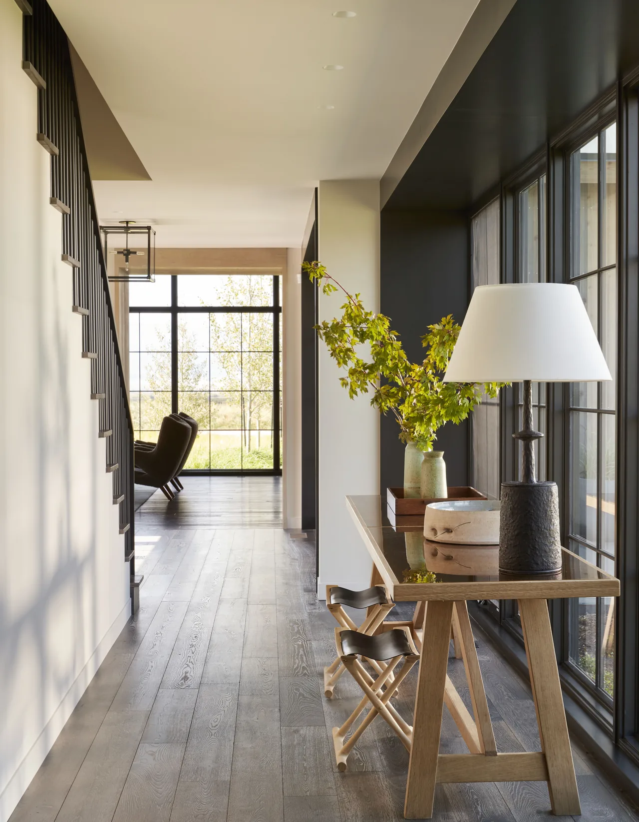 Interior view of a hallway featuring large grid windows and a wooden console table with a lamp and decorative greenery.
