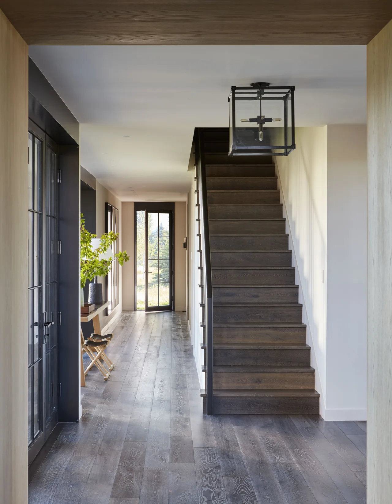 A modern hallway features dark wood stairs and flooring with black window and door frames letting in natural light.