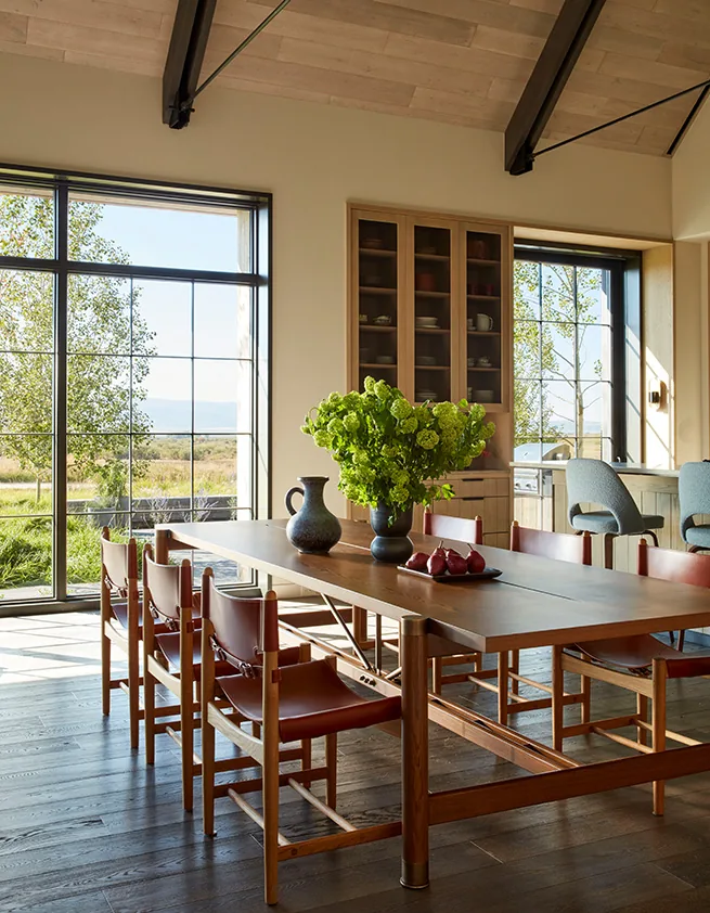 A dining space with leather strap chairs and a wooden table illuminated by natural light through large grid windows.