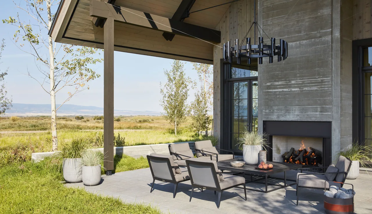Covered patio features modern outdoor furniture centered around a fireplace with a concrete surround and wood columns.