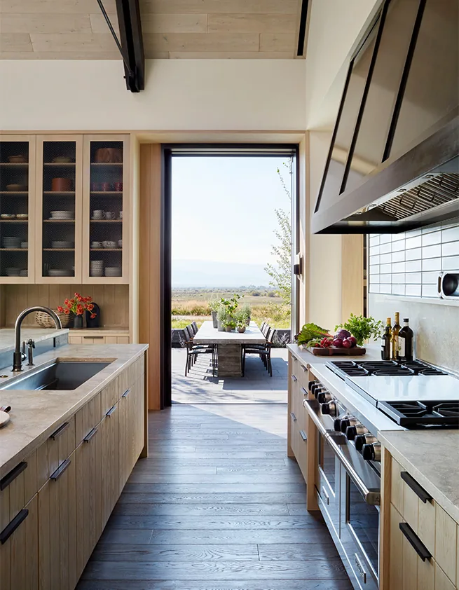Modern kitchen with wood cabinetry and a metal range hood opening to an outdoor dining patio with natural views.