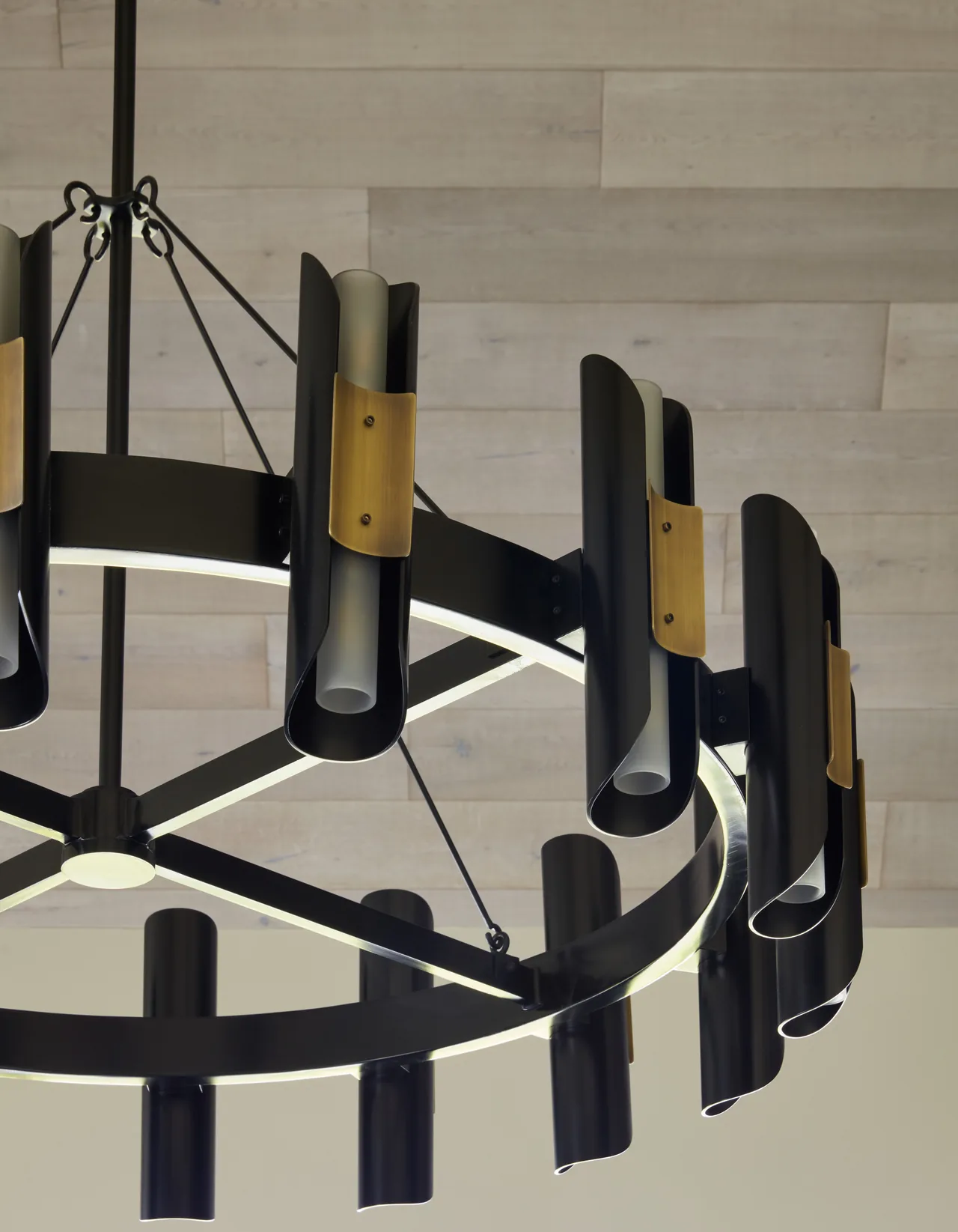 Modern circular chandelier features black metal, brass accents, and frosted glass tubes, hanging from a light wood-paneled ceiling.