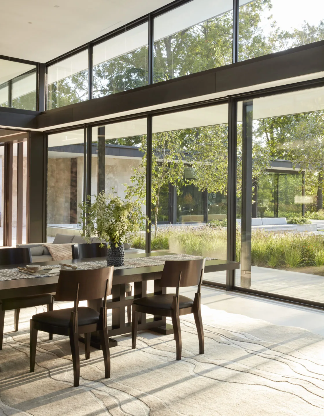 Dining area with dark wood table and chairs beside expansive floor-to-ceiling glass walls overlooking the garden.