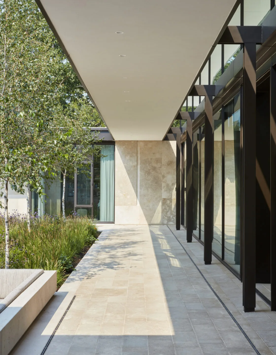 Covered exterior walkway with glass wall and black structural framing overlooking landscaped garden.