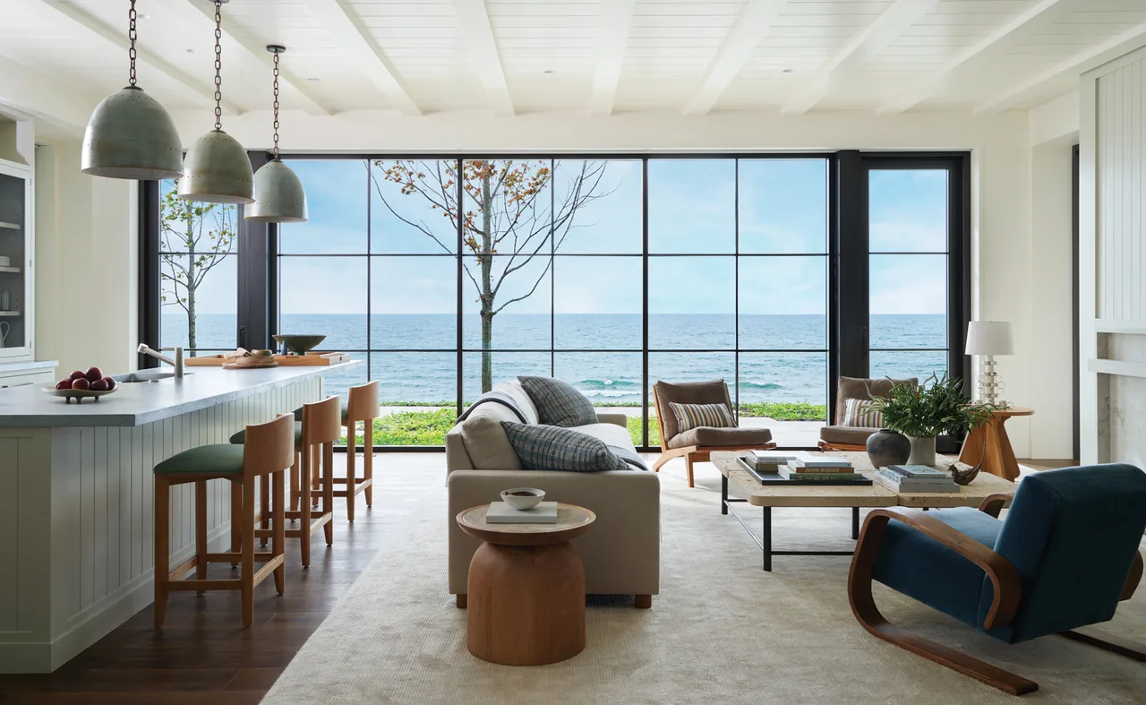 Living room and kitchen with black window frames overlooking the ocean, featuring wood bar stools and modern pendant lights.