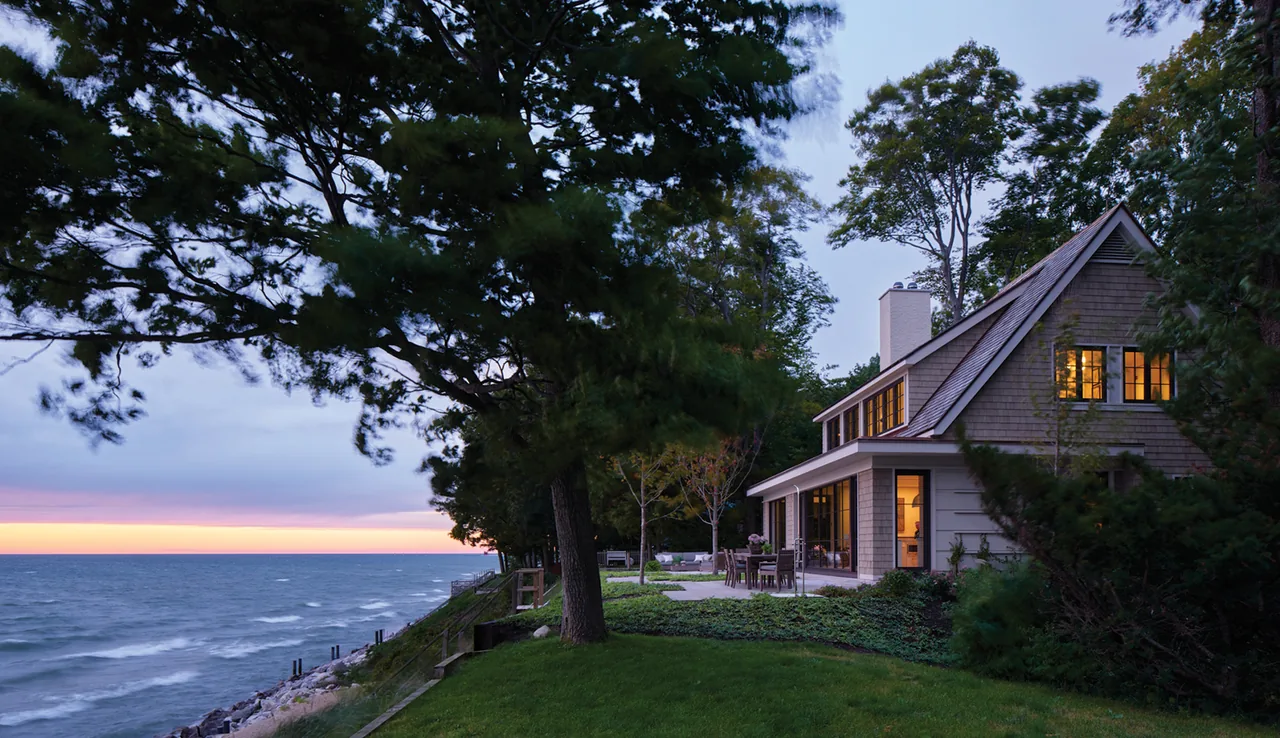 Lakeside house with shingle siding and a gable roof featuring multiple windows illuminated at dusk.