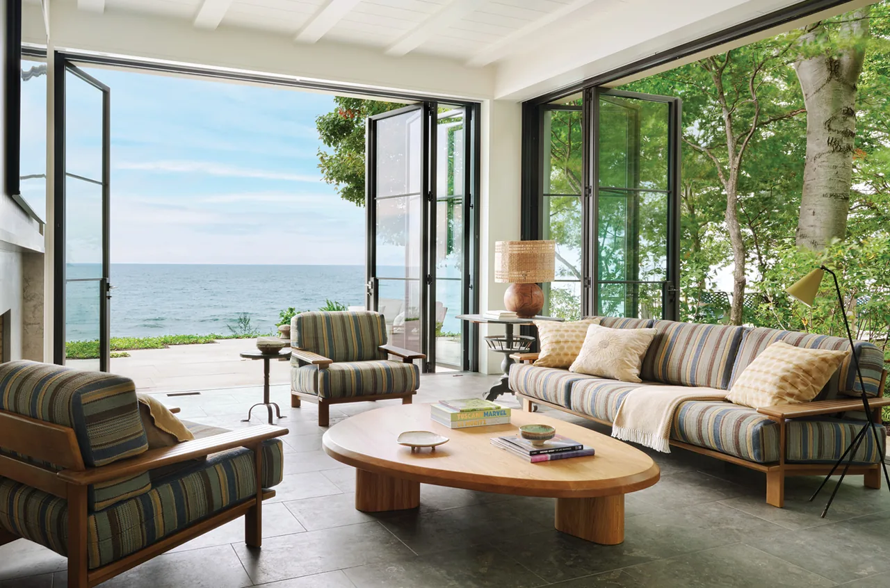Living room with wood frame furniture and folding black-framed glass doors opening to ocean and landscape views in project Making Waves.