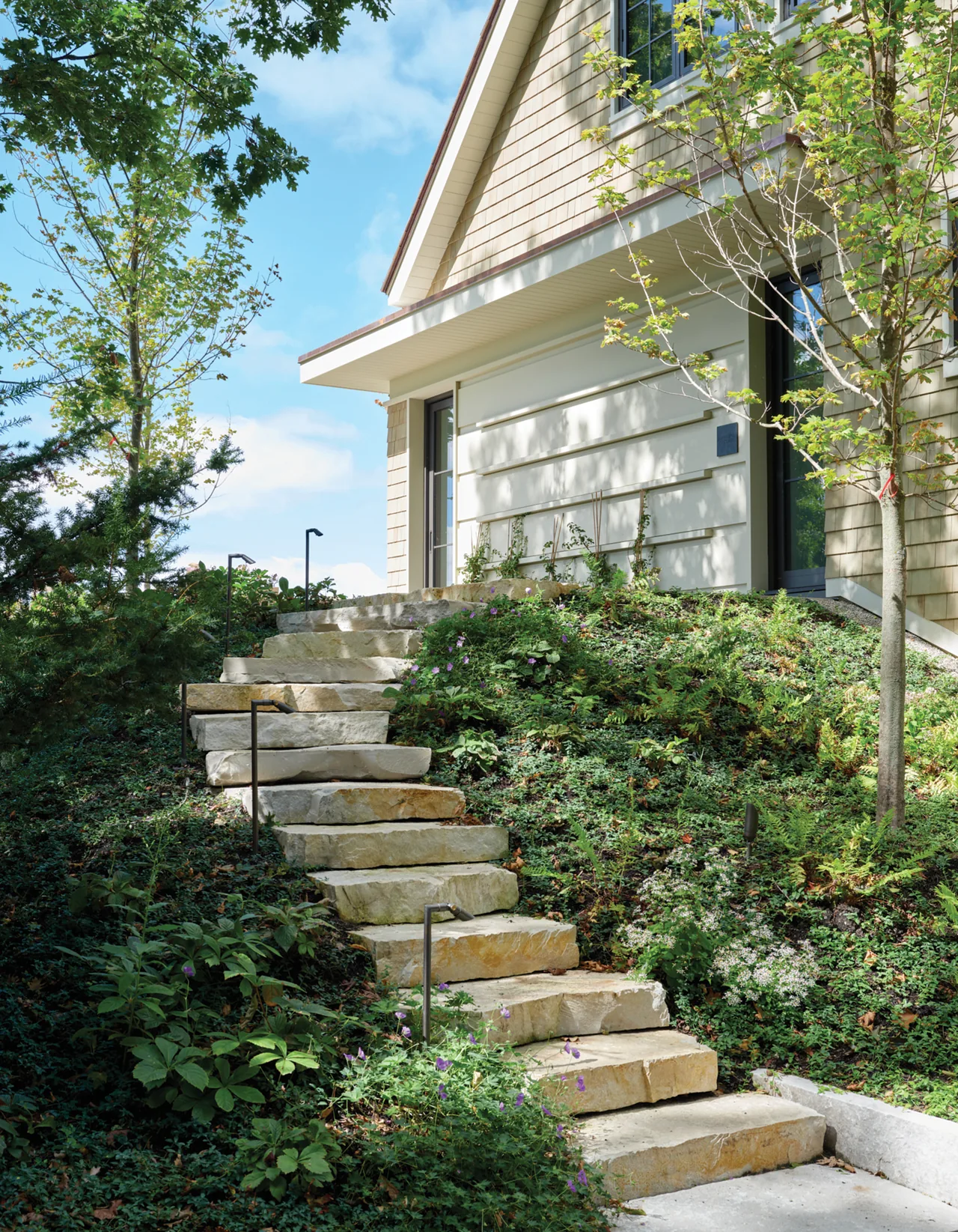 Exterior stone stairway ascends to a house with shingle siding and black window frames surrounded by natural landscaping.