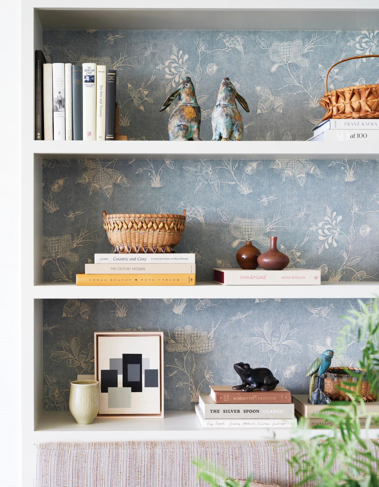 Built-in white bookshelf featuring decorative ceramics, books, and artistic decor with blue floral wallpaper background.